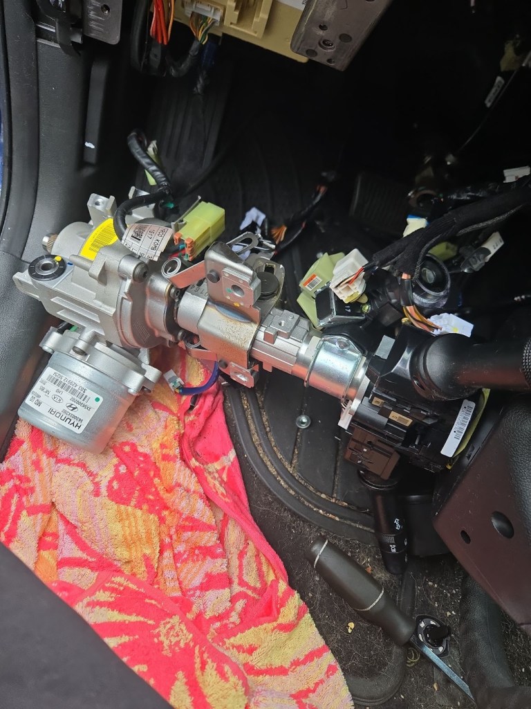 Close-up view of disassembled car steering column components, including an electric motor, wiring harness, and various connectors, resting on a patterned cloth inside a vehicle.