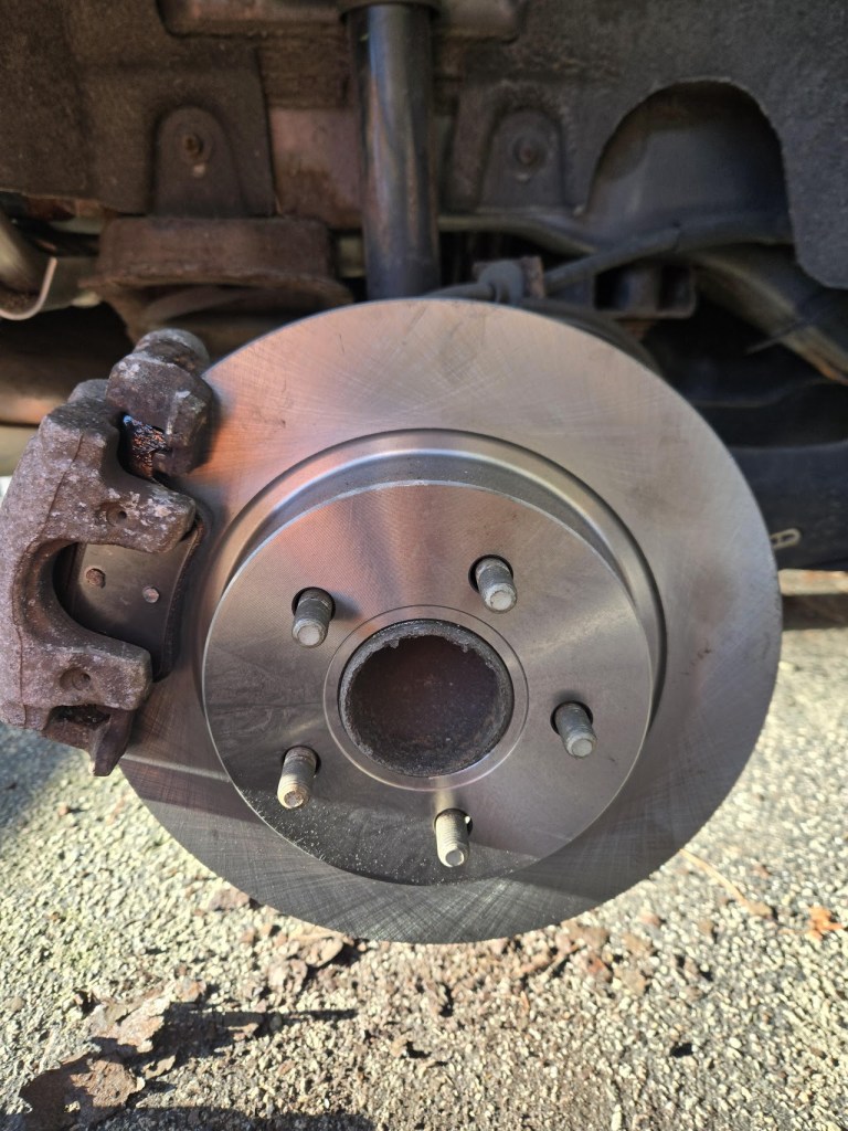 Close-up view of a car brake disc assembly showing the disc, bolts, and brake caliper against a textured surface.